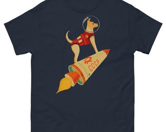 以下が含まれることがあります： ダークネイビーの半袖Tシャツ。宇宙ヘルメットと鎌と槌のエンブレムが付いた赤いベストを着用した茶色の犬のグラフィックが描かれています。犬はCCCPの文字と鎌と槌のシンボルがプリントされたロケットの上に立っています。