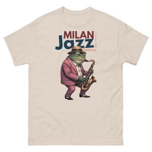 Puede incluir: Una camiseta beige de manga corta con el gráfico de una rana verde con un sombrero fedora marrón y un traje de rayas rosas mientras toca el saxofón. El texto dice MILAN Jazz FESTIVAL 11-14 May 1965 en letras azules, rojas y marrones.