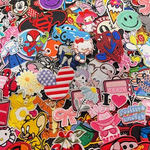 May include: A vibrant assortment of embroidered patches featuring cartoon characters, smiley faces, flowers, and patriotic designs. The patches include characters like Mickey Mouse, Spider-Man, and Hello Kitty, in various colors and shapes.