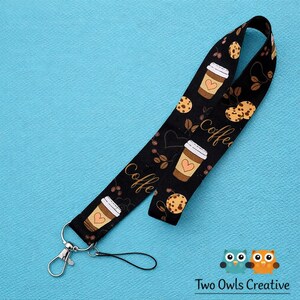May include: Black lanyard with a coffee and cookie pattern. The design includes coffee cups with heart accents, cookies, coffee beans, and the word "coffee" in a script font. It has a silver-colored clasp and a black string for attaching to a phone or ID badge.
