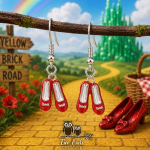 May include: A pair of silver-toned drop earrings featuring miniature red ballet slippers with small white bows. The earrings hang from silver hooks and are designed with a metallic finish, showcasing a vibrant red enamel coating on the shoe exterior.