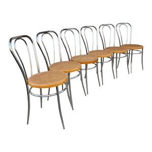 May include: A set of six vintage bistro chairs. Each chair features a woven rattan seat and a curved, chrome-finished metal frame. The chairs are arranged in a row, showcasing their classic design.