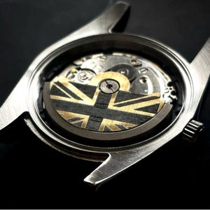 以下が含まれることがあります： 透明な裏蓋が付いたシルバーの腕時計のクローズアップ。複雑な機械式ムーブメントが見えます。ローターは黒と金のユニオンジャックのデザインです。時計の内部ギアとコンポーネントが見えます。