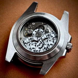 May include: A stainless steel watch case with a transparent back revealing the intricate mechanical movement. The watch face features a white background with a pattern of black skulls. The watch has a silver bezel and a crown on the side.