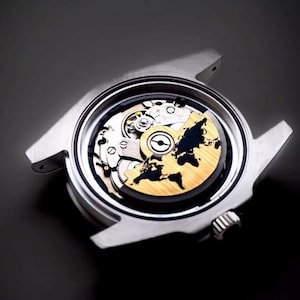 May include: A silver-toned wristwatch with a transparent face revealing the intricate inner workings. The watch face features a gold-colored world map design, with a black background. The watch has a sleek, modern design.