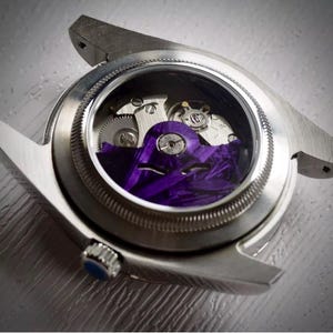 Puede incluir: Primer plano de un reloj de pulsera plateado con una parte trasera transparente, que revela los intrincados mecanismos internos. La esfera del reloj presenta un vibrante diseño púrpura. El reloj tiene una corona azul.
