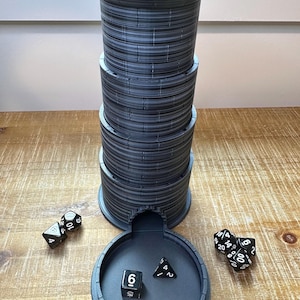 Puede incluir: Una torre de dados gris oscuro con diseño de castillo, con una base circular y una estructura cilíndrica alta. Varios dados en blanco y negro están esparcidos alrededor de la base, listos para un juego de mesa.