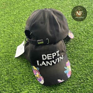 Puede incluir: Dos gorras de béisbol negras apiladas una encima de la otra. La parte delantera de la gorra inferior tiene el texto "DEPT. LANVIN" en blanco. La visera de la gorra inferior tiene un diseño floral colorido. Las gorras están sobre una superficie verde.