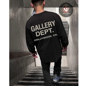 May include: Black long-sleeve shirt with the text "GALLERY DEPT. HOLLYWOOD, CA." in white. The shirt is worn by a person walking down a flight of stairs. The shirt has a crew neck and a relaxed fit.