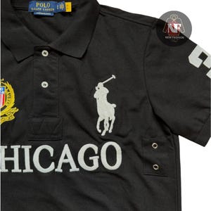 May include: Black polo shirt with white embroidered logo of a polo player on a horse. The shirt features the word "CHICAGO" in white block letters and a gold and red emblem. The shirt has a classic collar and short sleeves.