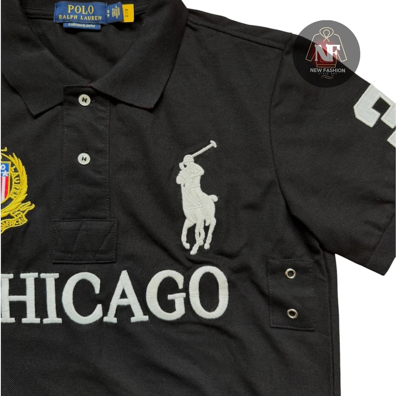 May include: Black polo shirt with white embroidered logo of a polo player on a horse. The shirt features the word "CHICAGO" in white block letters and a gold and red emblem. The shirt has a classic collar and short sleeves.
