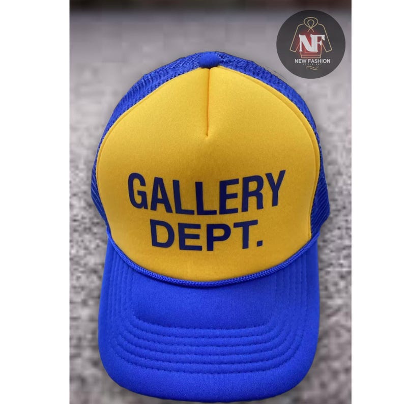 May include: A blue and yellow trucker hat with the text "GALLERY DEPT." in blue block letters. The hat has a mesh back and a curved brim. The hat is a classic style.
