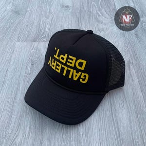 Puede incluir: Gorra de camionero negra con un logotipo 'GALLERY DEPT.' amarillo.