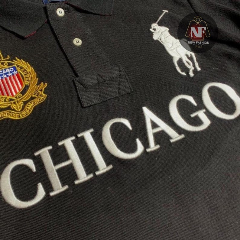 May include: Black polo shirt with white embroidered "CHICAGO" lettering across the chest. Features a white polo player logo and a crest with red, white, and blue accents. The shirt has a classic collar and button placket.