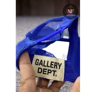 May include: A blue trucker hat with a mesh back and a curved brim. The hat features the text "GALLERY DEPT." on a rectangular patch. The hat has a snap closure for adjustable sizing.