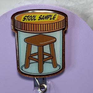 May include: A badge reel featuring a cartoon image of a stool inside a container, with the words "STOOL SAMPLE" on top. The reel has a clear plastic strap and a metal clip for attachment.