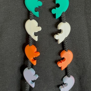 May include: Two black braided hoodie drawstrings featuring four pairs of colorful, heart-shaped puzzle piece beads in teal, white, orange, and light purple. The beads are threaded onto the cords, creating a symmetrical arrangement of matching halves.