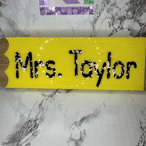 May include: A yellow pencil-shaped sign with the name "Mrs. Taylor" in black, dotted letters. The sign has a wooden end and is likely a teacher's nameplate or classroom decoration.