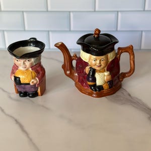 Könnte beinhalten: Ein Keramik-Tee- und Sahne-Set. Die Teekanne ist braun mit einem schwarzen Deckel und Griff, und der Sahneaufbereiter hat ein passendes Design. Beide Teile sind wie ein Mann mit Hut und Mantel geformt, der ein Getränk und Essen hält.