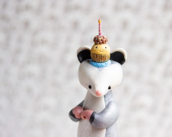 May include: A handcrafted figurine of an opossum wearing a blue scarf, with a cupcake on its head. The cupcake has a smiling face, chocolate sprinkles, and a lit candle. The opossum has gray and white fur, pink paws, and black ears.