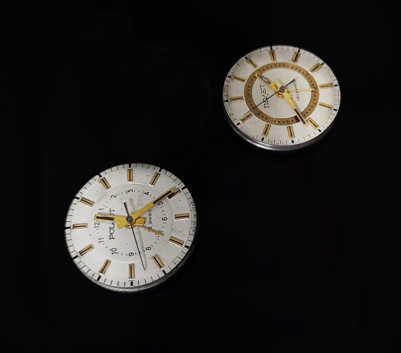 May include: Two vintage watch faces with white dials, gold hands, and gold hour markers. The watch faces have the word "POLJOT" printed on them. The background is black.