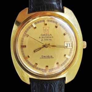 Op de afbeelding: Een goudkleurig Omega Genève Electronic f 300 Hz chronometer polshorloge met een ronde gouden wijzerplaat, streepindexen, een datumvenster op de drie-uurpositie en een zwarte, gestructureerde leren band. De wijzerplaat bevat het Omega-logo.