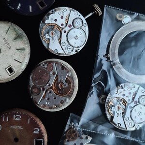 K&ouml;nnte beinhalten: Eine Sammlung von Vintage-Uhrenteilen, darunter Zifferbl&auml;tter und komplizierte mechanische Uhrwerke. Die Zifferbl&auml;tter weisen Gebrauchsspuren auf, mit dem Text "Wostok" sichtbar. Die Uhrwerke zeigen Zahnr&auml;der, Federn und andere kleine Komponenten.