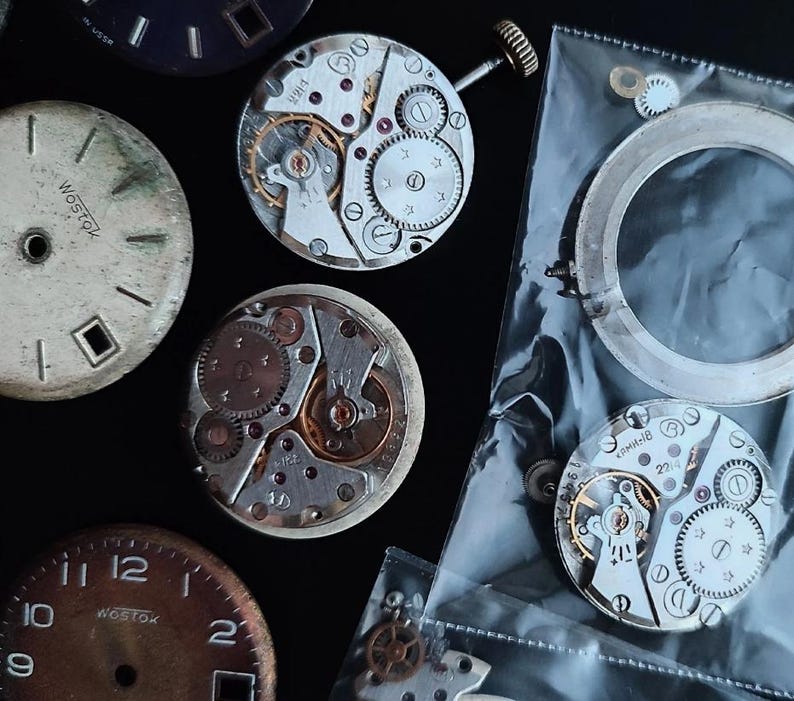 K&ouml;nnte beinhalten: Eine Sammlung von Vintage-Uhrenteilen, darunter Zifferbl&auml;tter und komplizierte mechanische Uhrwerke. Die Zifferbl&auml;tter weisen Gebrauchsspuren auf, mit dem Text "Wostok" sichtbar. Die Uhrwerke zeigen Zahnr&auml;der, Federn und andere kleine Komponenten.