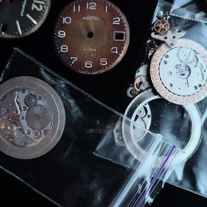 K&ouml;nnte beinhalten: Eine Sammlung von Vintage-Uhrenteilen, darunter Zifferbl&auml;tter und Innenmechanismen. Die Zifferbl&auml;tter sind schwarz und braun mit wei&szlig;en Ziffern. Einige Teile befinden sich in transparenten Plastikt&uuml;ten. Die Aufnahme erfolgt von oben.