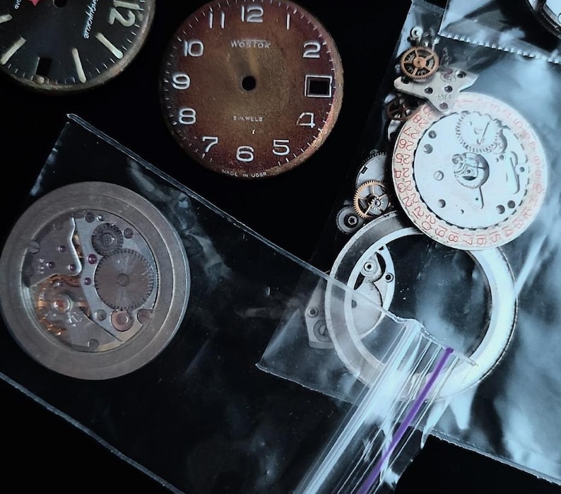 K&ouml;nnte beinhalten: Eine Sammlung von Vintage-Uhrenteilen, darunter Zifferbl&auml;tter und Innenmechanismen. Die Zifferbl&auml;tter sind schwarz und braun mit wei&szlig;en Ziffern. Einige Teile befinden sich in transparenten Plastikt&uuml;ten. Die Aufnahme erfolgt von oben.
