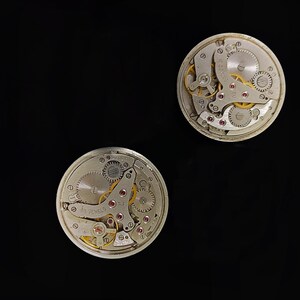 May include: Two circular vintage watch movements, showcasing intricate internal mechanisms. The silver-toned components include visible gears, springs, and red jewels, with gold accents. One movement displays the text "16 JEWELS". The image is set against a black backdrop.