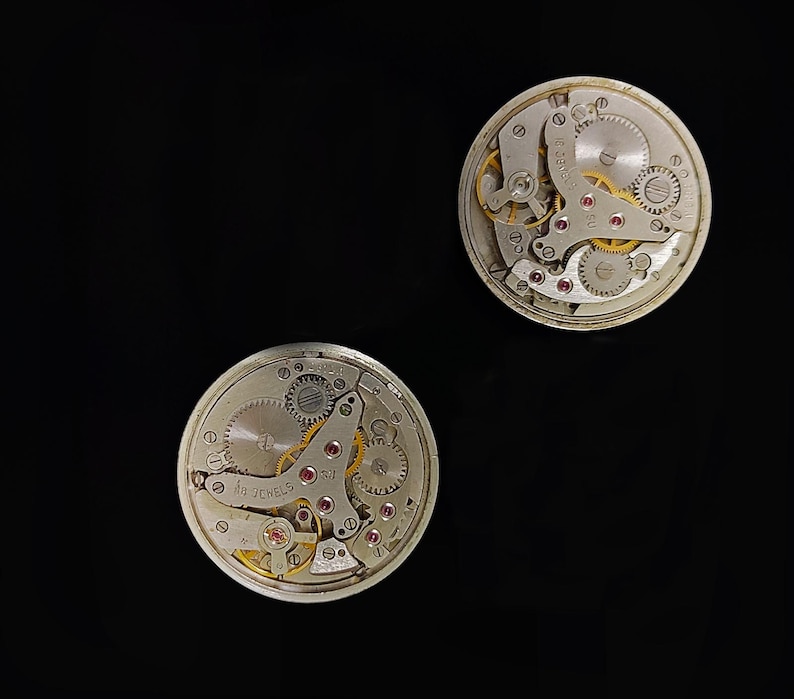 May include: Two circular vintage watch movements, showcasing intricate internal mechanisms. The silver-toned components include visible gears, springs, and red jewels, with gold accents. One movement displays the text "16 JEWELS". The image is set against a black backdrop.
