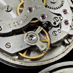 May include: Detailed view of a watch movement, revealing intricate gears, screws, and jewels. The metal parts are silver and gold, with red jewels. The text "18 JEWELS" is visible. The image highlights the complex inner workings of a mechanical watch.