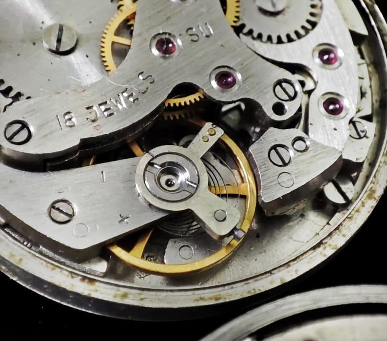 May include: Detailed view of a watch movement, revealing intricate gears, screws, and jewels. The metal parts are silver and gold, with red jewels. The text "18 JEWELS" is visible. The image highlights the complex inner workings of a mechanical watch.