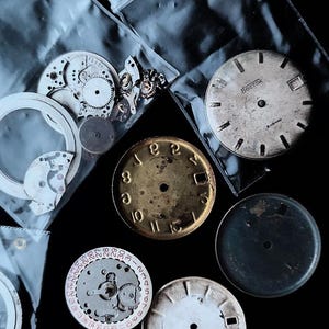 K&ouml;nnte beinhalten: Eine Sammlung von Vintage-Uhrenteilen, darunter Zifferbl&auml;tter, Zahnr&auml;der und Innenmechanismen, auf dunklem Untergrund angeordnet. Die Zifferbl&auml;tter zeigen r&ouml;mische und arabische Ziffern mit verschiedenen Farben und Texturen.