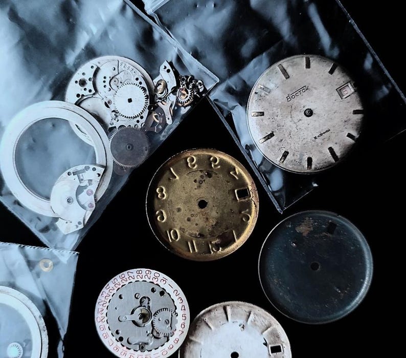 K&ouml;nnte beinhalten: Eine Sammlung von Vintage-Uhrenteilen, darunter Zifferbl&auml;tter, Zahnr&auml;der und Innenmechanismen, auf dunklem Untergrund angeordnet. Die Zifferbl&auml;tter zeigen r&ouml;mische und arabische Ziffern mit verschiedenen Farben und Texturen.