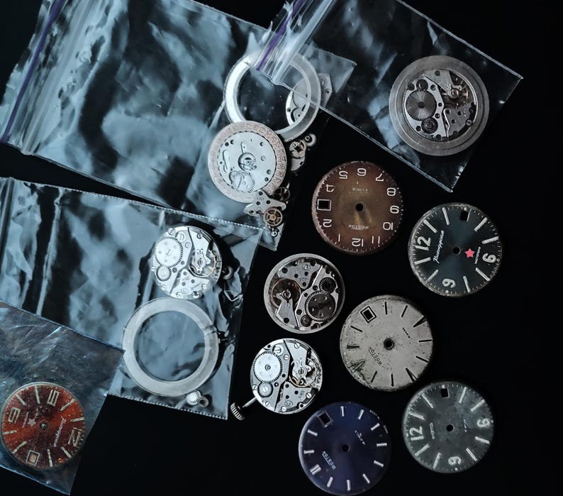 K&ouml;nnte beinhalten: Eine Sammlung von Vintage-Uhrenteilen, darunter Zifferbl&auml;tter mit Ziffern und Zeigern sowie zerlegte Uhrwerkkomponenten. Einige Teile befinden sich in transparenten Plastikt&uuml;ten. Die Zifferbl&auml;tter sind in verschiedenen Farben erh&auml;ltlich, darunter Braun, Schwarz und Blau.