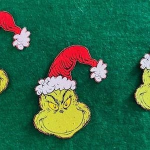 May include: Three Grinch-themed cutouts. Each cutout features the Grinch's face in yellow-green with a red Santa hat trimmed with white fur. The cutouts are arranged on a green background.