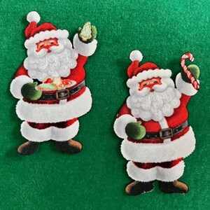 May include: Two felt Christmas ornaments of Santa Claus. One Santa is holding a plate of food and a green tree. The other Santa is holding a candy cane.