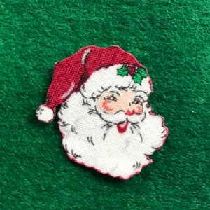 May include: A red and white felt appliqué of Santa Claus's face with a white beard and a red hat with green holly.