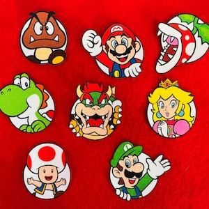 May include: A collection of eight circular pins featuring colourful cartoon characters including a brown Goomba, Mario, a green Piranha Plant, a green Yoshi, Bowser, Princess Peach, a red and white Toad, and Luigi on a bright red background.