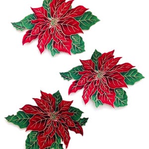 May include: Three decorative poinsettia flowers with red petals and green leaves. Each flower has gold-colored accents and is set against a white background. These festive floral decorations are perfect for holiday decor.