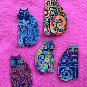 May include: Five decorative cat shapes with vibrant designs. Each cat features unique patterns of flowers, swirls, and stripes in shades of blue, purple, orange, and green. Gold outlines are set against a pink background.