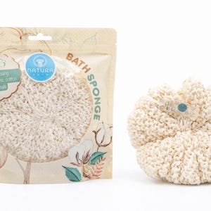 May include: A natural-colored bath sponge made from organic cotton, with a button detail. The sponge is next to its packaging, which states "Crafted using 100% organic cotton materials."