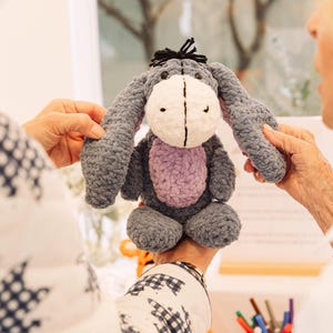 May include: A gray and purple crocheted stuffed donkey toy with black ears and a black mane. The donkey is being held by a person's hands.