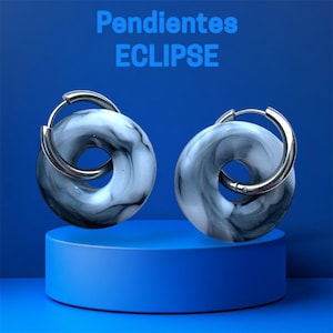 Puede incluir: Un par de pendientes Eclipse con un diseño veteado gris y blanco. Los pendientes son circulares con un cierre de aro plateado. El texto "Pendientes ECLIPSE" se muestra en azul en la parte superior de la imagen. Los pendientes se exhiben sobre un pedestal azul.