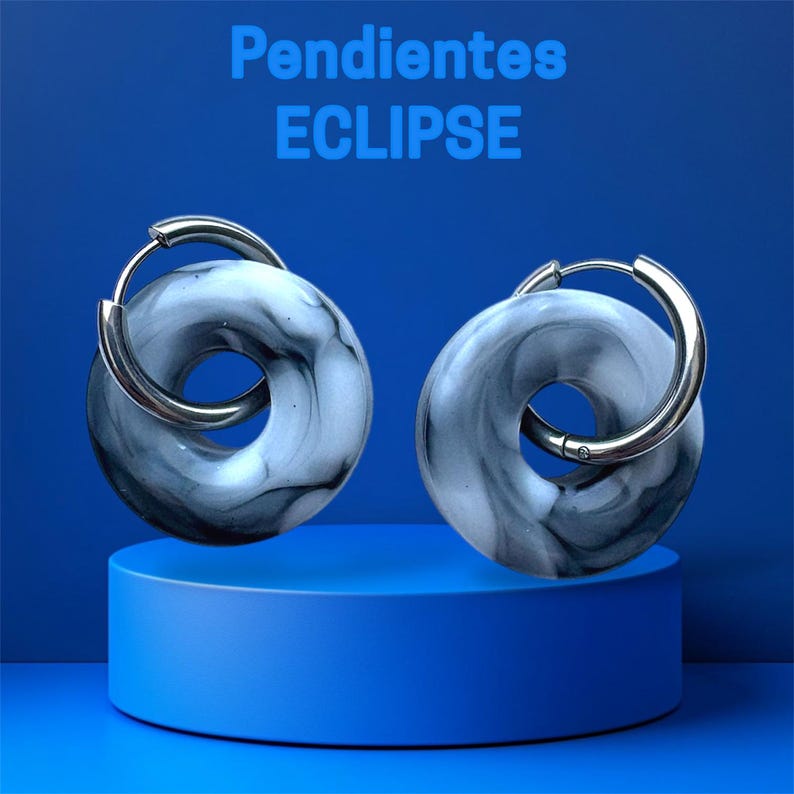 Pode incluir: Um par de brincos Eclipse com um design marmorizado cinza e branco. Os brincos s&atilde;o circulares com um fecho de argola prateada. O texto "Pendientes ECLIPSE" &eacute; exibido em azul na parte superior da imagem. Os brincos s&atilde;o exibidos em um pedestal azul.