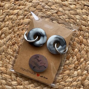 Pode incluir: Um par de brincos cinza e branco marmorizados com argolas prateadas, embalados em um cart&atilde;o marrom. O cart&atilde;o tem um adesivo que diz "Thank You" e "Handmade with love".
