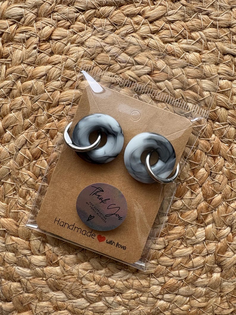 Pode incluir: Um par de brincos cinza e branco marmorizados com argolas prateadas, embalados em um cart&atilde;o marrom. O cart&atilde;o tem um adesivo que diz "Thank You" e "Handmade with love".