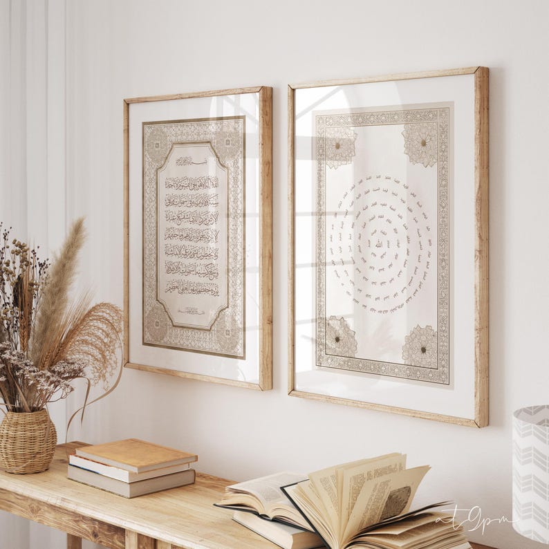 May include: Two framed Islamic art prints with gold and beige tones. The prints feature Arabic calligraphy and geometric designs, set in light wood frames. The artwork is displayed on a wooden table with books and decorative elements.